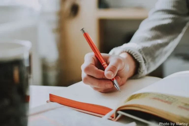 A person's hand holding an orange pen writing in a journal. Manifesting how to write better.