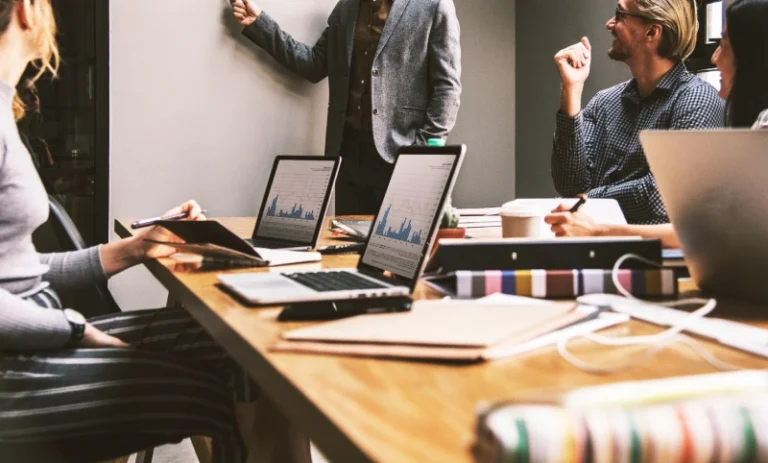 A group of people at work in a meeting. One person is standing up giving a presentation while others are seated and using their laptops. How to manifest a job.