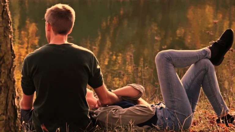 Guy sitting in grass with his back facing camera and his girlfriend laying down with her head in his lap. How to manifest a boyfriend.