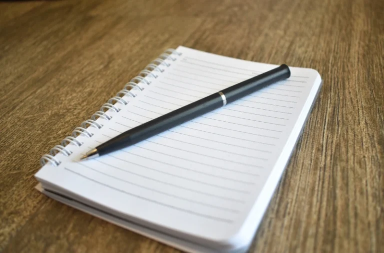 A notepad laying on a wooden table with a pen placed diagonally on top of it. How to manifest on paper.