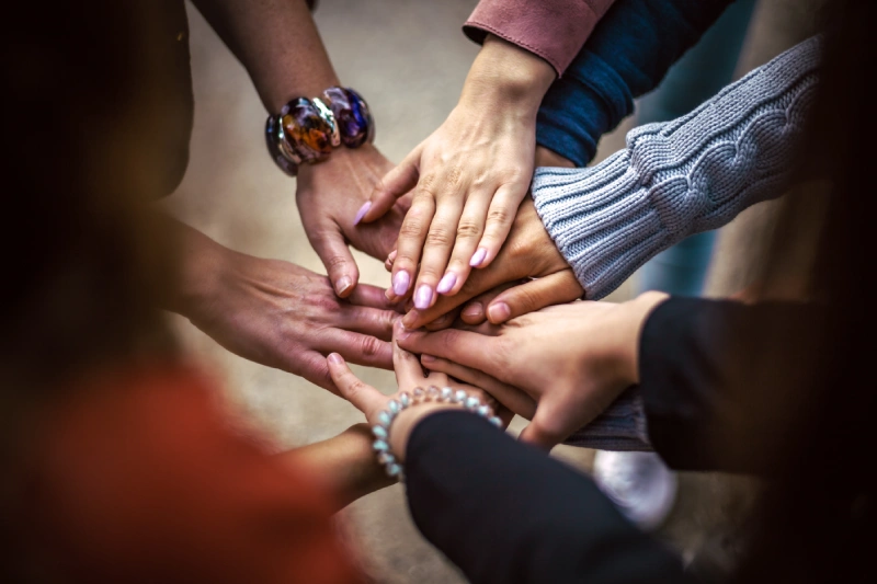 A group of individuals representing various nationalities, united in a circle with their hands stacked on top of each other. Collaborative manifestation.