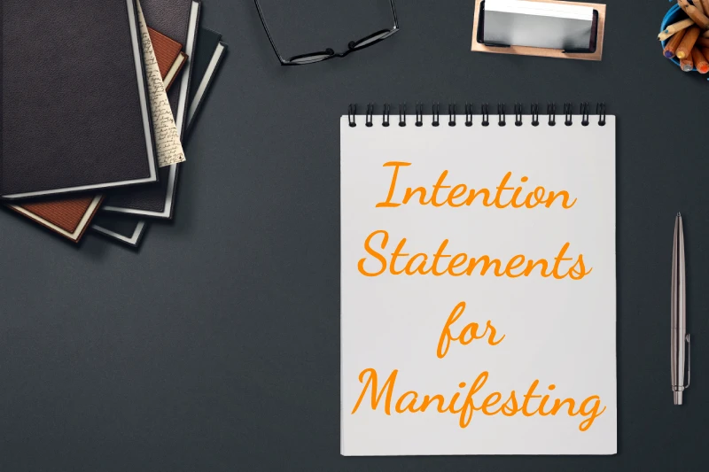 A notepad on a black background surrounded by office supplies. In script writing, the notepad reads "how to write intention statements for manifesting."